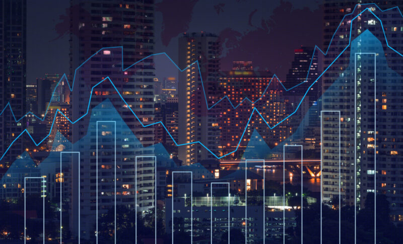 City skyline at night with illuminated high-rise buildings, overlaid with abstract financial graphs including bar charts and line graphs, representing data analytics, market trends or economic activity.