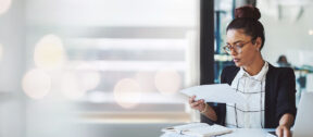 Businesswoman reviewing financial documents at a desk with charts, a notebook, and a laptop, focused in a modern office setting.