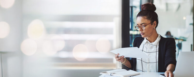 Businesswoman reviewing financial documents at a desk with charts, a notebook, and a laptop, focused in a modern office setting.