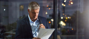 A business professional stands by a window at night, reviewing printed financial documents with glasses in hand, reflecting the considered work behind a financial audit.