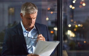 A business professional stands by a window at night, reviewing printed financial documents with glasses in hand, reflecting the considered work behind a financial audit.