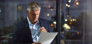 A business professional stands by a window at night, reviewing printed financial documents with glasses in hand, reflecting the considered work behind a financial audit.