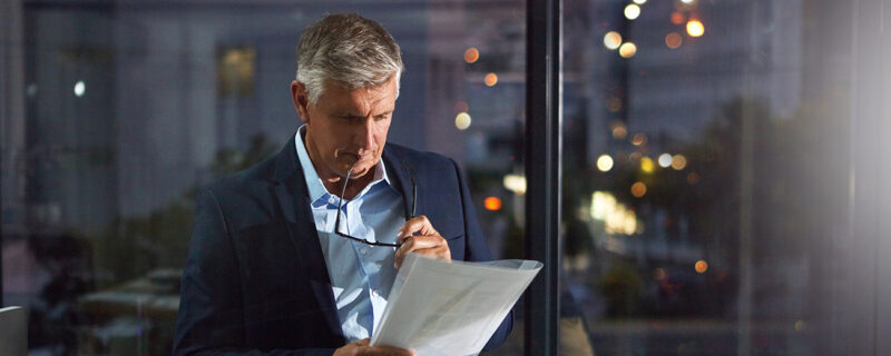 A business professional stands by a window at night, reviewing printed financial documents with glasses in hand, reflecting the considered work behind a financial audit.