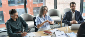 A group of company directors seated around a boardroom table in a modern office, reviewing printed reports, charts and laptops together during a considered governance discussion.