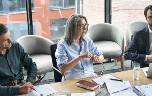 A group of company directors seated around a boardroom table in a modern office, reviewing printed reports, charts and laptops together during a considered governance discussion.