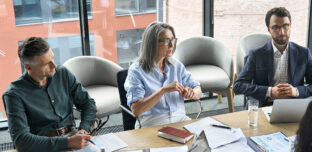 A group of company directors seated around a boardroom table in a modern office, reviewing printed reports, charts and laptops together during a considered governance discussion.