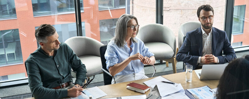 A group of company directors seated around a boardroom table in a modern office, reviewing printed reports, charts and laptops together during a considered governance discussion.