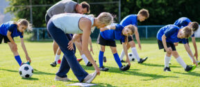 A group of children in blue kit stretch during a football training session on a grassy field, guided by two coaches in an outdoor community setting.