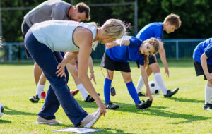 A group of children in blue kit stretch during a football training session on a grassy field, guided by two coaches in an outdoor community setting.