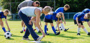 A group of children in blue kit stretch during a football training session on a grassy field, guided by two coaches in an outdoor community setting.