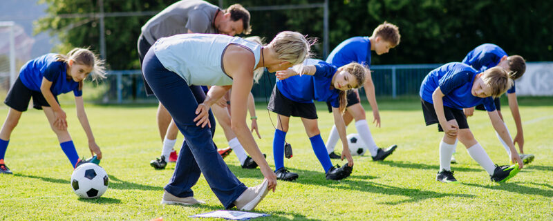 A group of children in blue kit stretch during a football training session on a grassy field, guided by two coaches in an outdoor community setting.