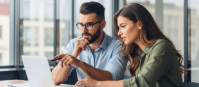 Two business colleagues sit together at a desk with financial documents, looking at a laptop as they discuss options for their trust structure.