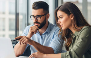 Two business colleagues sit together at a desk with financial documents, looking at a laptop as they discuss options for their trust structure.