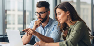 Two business colleagues sit together at a desk with financial documents, looking at a laptop as they discuss options for their trust structure.