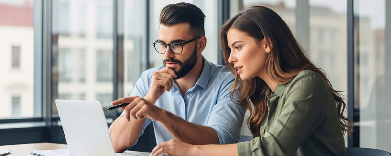 Two business colleagues sit together at a desk with financial documents, looking at a laptop as they discuss options for their trust structure.
