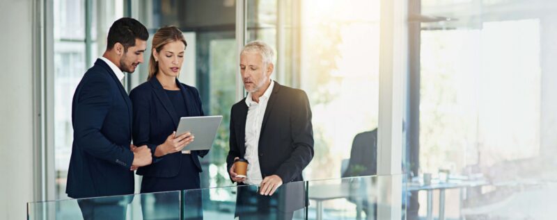 Three colleagues standing together in a bright modern office, reviewing information on a tablet during a discussion.