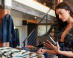 A retail worker checks stock on a tablet beside a table of folded jumpers in a warmly lit clothing store with a timber mezzanine.