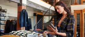 A retail worker checks stock on a tablet beside a table of folded jumpers in a warmly lit clothing store with a timber mezzanine.