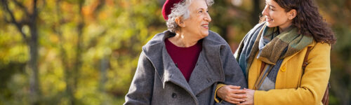 An older person in a red beret and grey coat walks arm in arm with a younger person in a yellow coat and checked scarf through an autumn park.